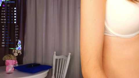 Media: Video of a woman's arm in a white bra, viewed from the side, in a dimly lit room with a table, chair, and pink flowers.