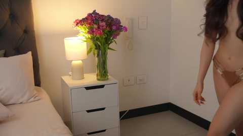 Media: Video of a nude woman with long dark hair standing near a white dresser with a lamp and flowers, in a minimalist bedroom with beige walls and black baseboards.