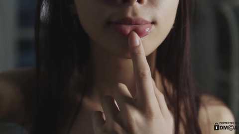 Media: A close-up video of a young woman with medium brown skin, long black hair, and full lips. She's touching her lips with her index finger, wearing a strapless top. Background is blurred.