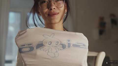 Media: Video of a woman with glasses, medium build, wearing a white \"Barbie\" T-shirt featuring a cartoonish Barbie logo. Background shows a blurred, bright room with white walls and a window.