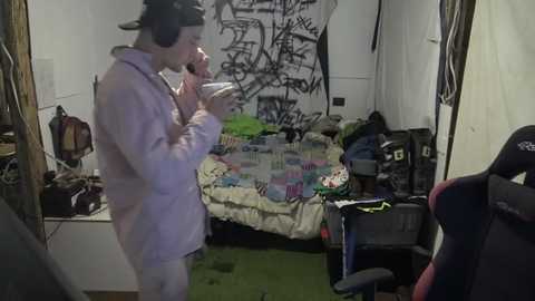 Media: Video of a cluttered bedroom with a man in a white hoodie and black cap, speaking on a phone, surrounded by messy clothing and a graffiti-covered wall.