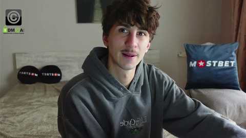 Media: Video of a young man with tousled brown hair, wearing a gray hoodie, smiling in a bedroom with a bed, pillows, and a TV in the background.