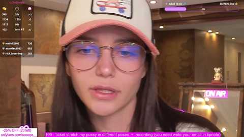 Media: A video of a young woman with fair skin and long dark hair, wearing a white cap with pink trim and round glasses, in a cozy, dimly lit room with wooden decor.