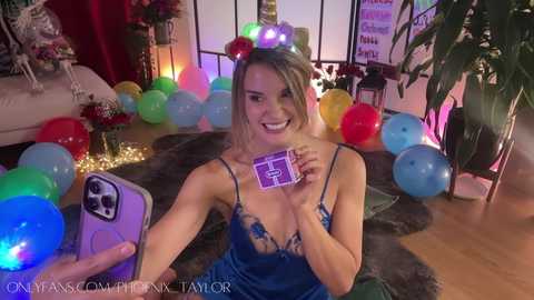 Media: Video of a smiling, blonde, light-skinned woman in a blue lace lingerie top, surrounded by colorful balloons, holding a purple box, in a festive, dimly lit room.