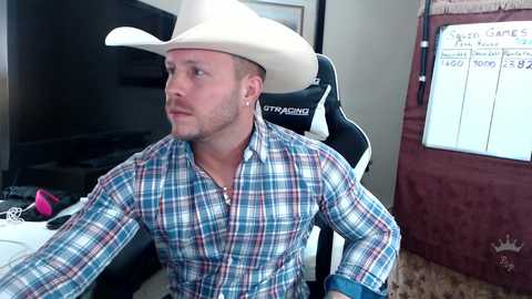 Media: Video of a muscular, light-skinned man with short hair and a beard, wearing a plaid shirt and white cowboy hat, seated in a gaming chair, in a dimly lit room with a TV and gaming setup.