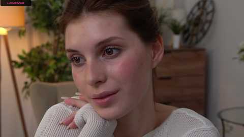 Media: Video of a fair-skinned woman with brown hair in a white knit sweater, blushing slightly, sitting indoors with plants and wooden furniture in the background.