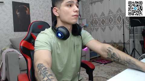 Media: Video of a young, muscular, tattooed man with short hair, wearing a green polo shirt, playing video games on a red and black gaming chair in a room with white floral wallpaper and a QR code watermark.