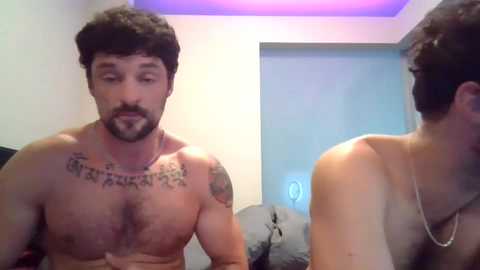 Media: Video of a shirtless, muscular man with a beard and tattoos, including a large one on his chest, standing in a dimly lit bedroom with a gray bed and a window.