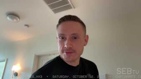 Media: Video of a young, clean-shaven man with short brown hair, wearing a black T-shirt, indoors, with a ceiling vent and blurred background.