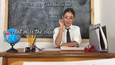 Media: Video of a young woman in glasses, wearing a white shirt and striped tie, sitting at a wooden desk in a classroom.