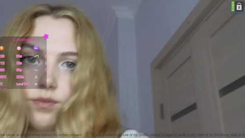 Media: Video of a young woman with blonde wavy hair, light skin, and blue eyes, partially obscured by a pink overlay displaying \"Live\" and chat options. She's indoors, next to a white door, with a white wall background.