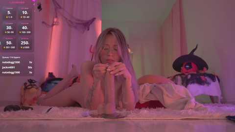 Media: A video of a nude, slender woman with long blonde hair lying on a white shag rug in a dimly lit, whimsical room with a plush toy and pink drapes.