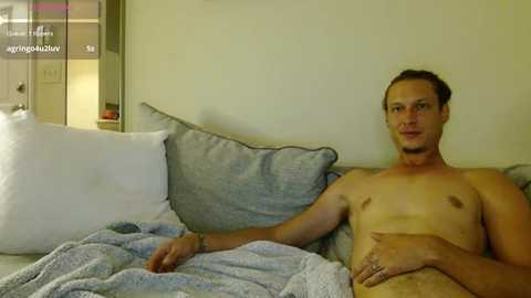 Media: A video of a shirtless, fair-skinned, slim-bodied man with short hair, reclining on a gray couch under a light gray blanket, surrounded by white pillows, in a dimly lit room.