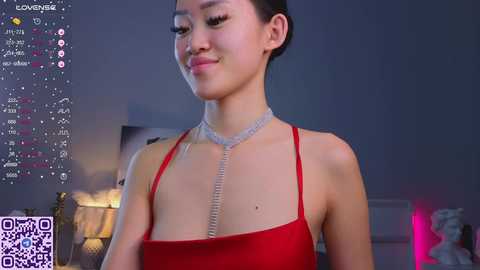 Media: Video of a young Asian woman with fair skin, dark hair, and a slender build, wearing a red halter dress with a silver chain necklace, smiling. Background shows a dimly lit room with a lamp and a bed.