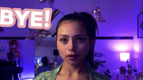 Media: Video of a young woman with light skin, dark hair in a high ponytail, wearing a green top, in a dimly lit room with purple lighting, butterflies on the wall, and a \"BIE!\" sign.