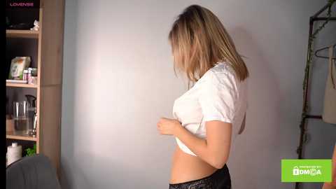 Media: Video of a light-skinned woman with shoulder-length blonde hair, lifting a white t-shirt to reveal her midriff and black lace panties. Background includes a wooden shelf and a green logo.