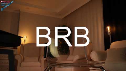 Media: Video of a dimly lit, modern living room with beige walls, white armchairs, a glass coffee table, and a crescent moon-shaped mirror. The text \"BBR\" is overlaid on the image in white.