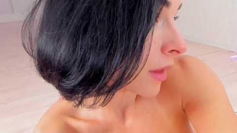 Media: A close-up video of a fair-skinned woman with shoulder-length, straight black hair, gazing to the right. She has a smooth complexion and is topless, suggesting a nude or partially nude state. The background is a plain, light-colored wall.