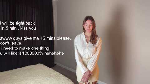 Media: Video of a young, fair-skinned woman with long brown hair, wearing a white shirt and standing in a bare room with beige walls. Text overlay: \"I will be right back in 5 min, kiss you, awww guys give me 15 mins please, don't leave, I need make one thing u will like it 1000000% hahahehe\