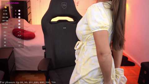 Media: Video of a woman in a yellow dress, standing beside a black gaming chair, in a bedroom with pink walls and a bed.