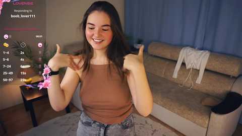 Media: Video of a young woman with long brown hair, wearing a brown tank top and blue jeans, posing in a living room with a beige sofa and blue curtains.