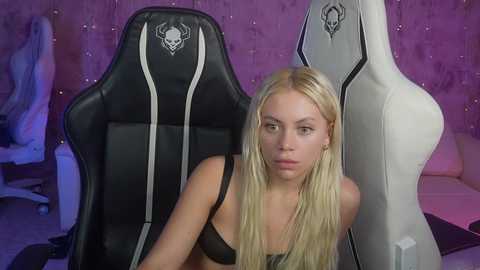 Media: Video of a blonde woman with fair skin and medium build, wearing a black bra, sitting in a gaming chair with an alien skull logo. Pink and purple wall background.