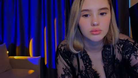 Media: Video of a young Caucasian woman with fair skin, straight blonde hair, and light pink lips, wearing a black lace robe. Background features dark curtains and a lit lamp.