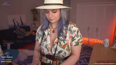 Media: Video of a plus-sized woman with long blue hair, wearing a white hat and floral blouse, standing in a dimly lit room with a blue sofa, plants, and neon \"CLEAN\" sign.