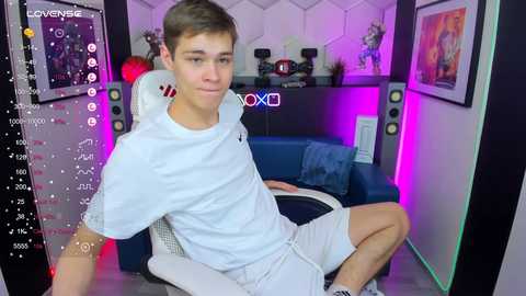 Media: Video of a teenage boy in a white t-shirt and shorts, sitting on a blue gaming chair in a dimly-lit room with purple and pink lights, surrounded by gaming equipment and framed posters.