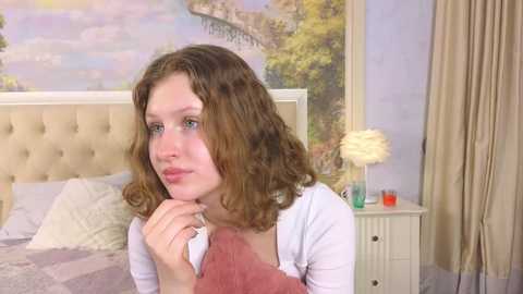 Media: A video of a young woman with curly brown hair, blue eyes, and pale skin, wearing a white cardigan, looking contemplative in a cozy bedroom with a beige tufted headboard, floral wallpaper, and a cream-colored chair.