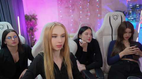 Media: Video of four young women, one with long blonde hair, seated in white gaming chairs, focused on their phones in a cozy, brightly lit room with pink and purple lights.