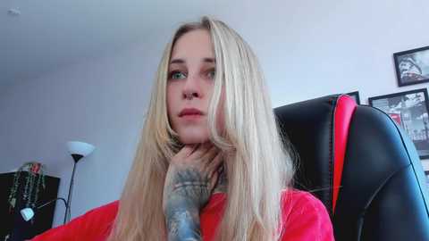 Media: Video of a blonde woman with tattoos on her neck, wearing a red top, sitting on a black chair, looking thoughtful. Background includes a lamp, framed pictures, and a red coat draped over the chair.