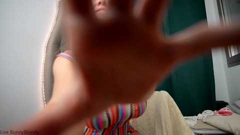 Media: A video of a woman with medium skin tone, wearing a colorful striped dress, standing in a bedroom with beige walls and green curtains. Her hands are raised in front of her face, blurring her features.