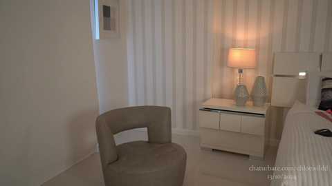 Media: A video of a minimalist bedroom with a beige armchair, white nightstand, lamp, and sheer white curtains.