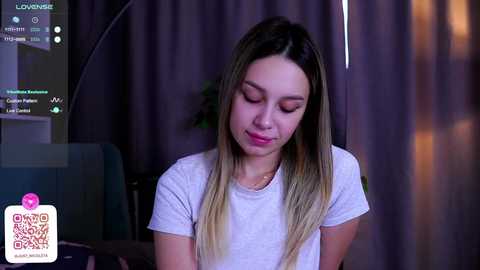 Media: Video of a young woman with long, straight blonde hair, light skin, wearing a white t-shirt, looking down, in a dimly lit room with dark curtains.