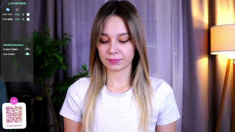 Media: Video of a young Caucasian woman with long, blonde hair, wearing a white t-shirt, smiling softly. Background includes a potted plant, a lamp, and purple curtains.