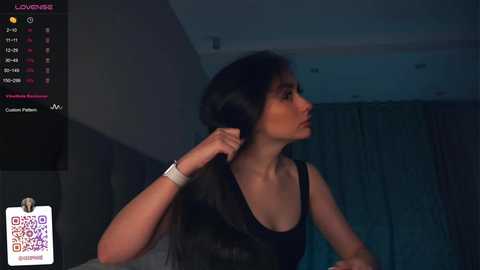 Media: A video of a young woman with long, dark hair, wearing a black tank top, adjusting her hair in a dimly lit room with curtains in the background.