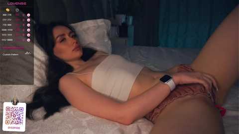 Media: Video of a woman with long black hair, fair skin, and wearing a white tank top and pink lace panties, lying on a bed, hand on her groin. Background shows dimly lit room with white curtains.