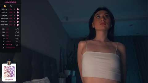 Media: Video of a slender woman with long black hair, wearing a white tank top, standing indoors at night, with a dimly lit room and a digital clock in the background.