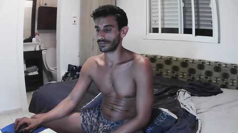 Media: Video of a shirtless South Asian man with short black hair, wearing patterned shorts, sitting on a bed in a messy room with a green-patterned headboard and white shutters.