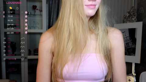 Media: Video of a blonde woman with long hair, wearing a strapless light pink top, in a dimly lit room with a white bookshelf and a framed picture in the background.