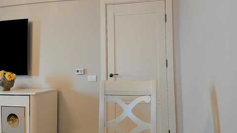 Media: A video of a minimalist, beige room featuring a white door with a cross design, a flat-screen TV on the left, a white dresser with a vase of yellow flowers, and a white chair.