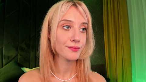 Media: Video of a young, fair-skinned woman with straight, shoulder-length blonde hair, wearing a white pearl necklace, sitting in front of a dark, patterned wall and green curtains. She has blue eyes and a slight smile.