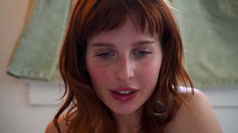 Media: A close-up video of a fair-skinned woman with long, wavy red hair and light makeup, looking slightly downwards with a neutral expression. The background shows a blurred, pale green wall.