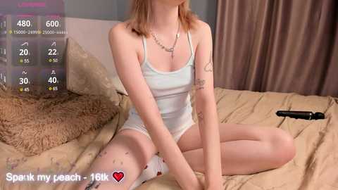 Media: A video of a young woman with pale skin and light brown hair, wearing a sheer white lingerie set, sitting on a beige bed. A black dildo is on the bed.