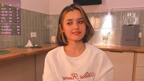 Media: Video of a young woman with light brown hair, wearing a white sweatshirt with \"Jasmine's Restaurant\" in red script, sitting in a modern kitchen with green tiled walls, wooden countertops, and a microwave.