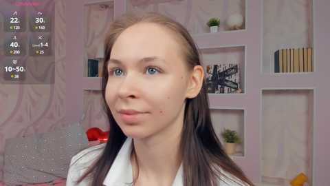 Media: Video of a fair-skinned woman with straight, light brown hair, blue eyes, and a white shirt, standing indoors in a pastel-colored room with bookshelves and a red heart-shaped pillow.