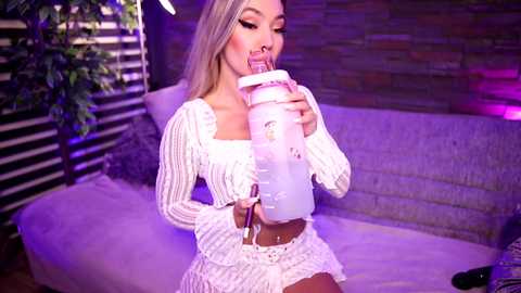 Media: Video of a blonde woman with long hair, wearing a white knitted crop top and short skirt, drinking from a baby bottle, against a purple-lit brick wall, potted plant, and bed.