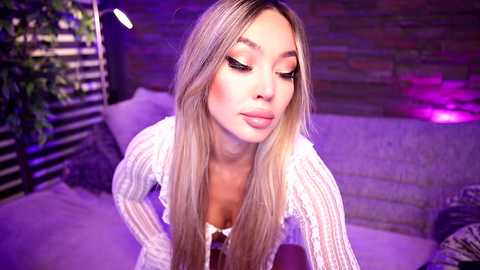 Media: Video of a young Asian woman with long blonde hair, wearing a white, ribbed sweater, with a soft, dimly lit, brick-walled background. She has makeup with heavy eyeliner and pink lipstick.