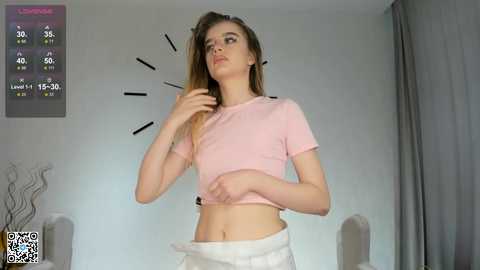 Media: Video of a young woman with long, straight brown hair, wearing a pink crop top and white pants, standing in a dimly lit room. A temperature gauge is visible on the left wall.